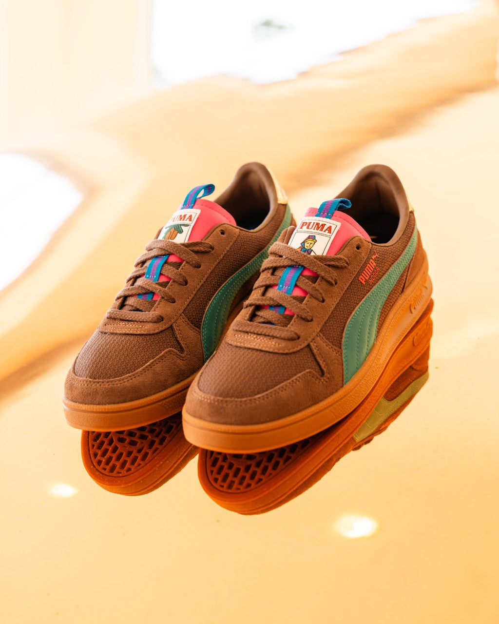 Ein Paar Rothaus Indoor Tannenzäpfle von Puma mit blauen Streifen, rosa Zungenetiketten und blauen Akzenten wird auf einer orangefarbenen, reflektierenden Oberfläche präsentiert.