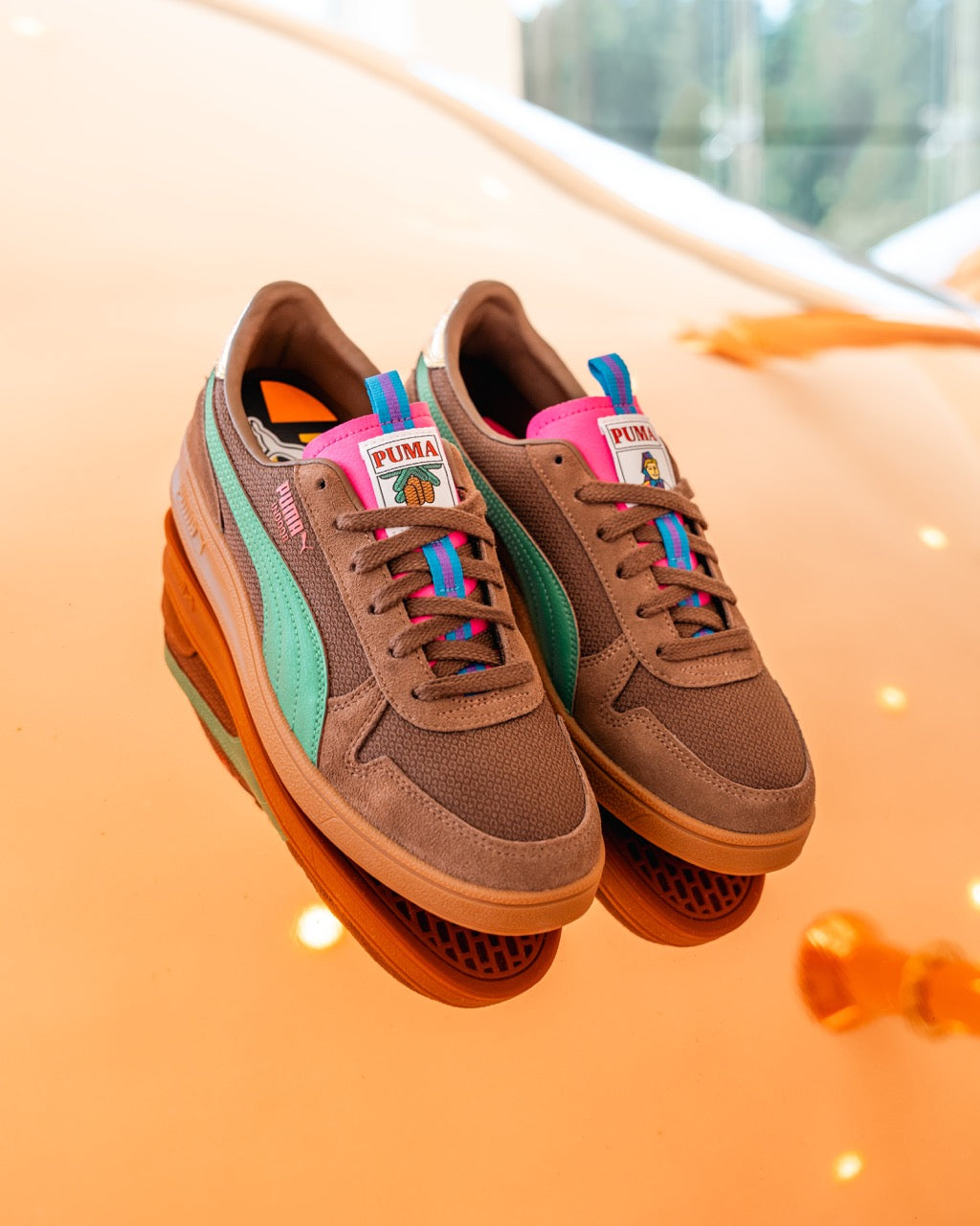 Die Rothaus Indoor Tannenzäpfle Sneakers von Puma haben ein braunes Obermaterial, blaugrüne Seitenstreifen, pinke und blaugrüne Akzente und mehrfarbige Schnürsenkel, die auf einer leuchtend orangefarbenen Oberfläche präsentiert werden und von Tannenzäpfle Kultbier inspiriert sind.