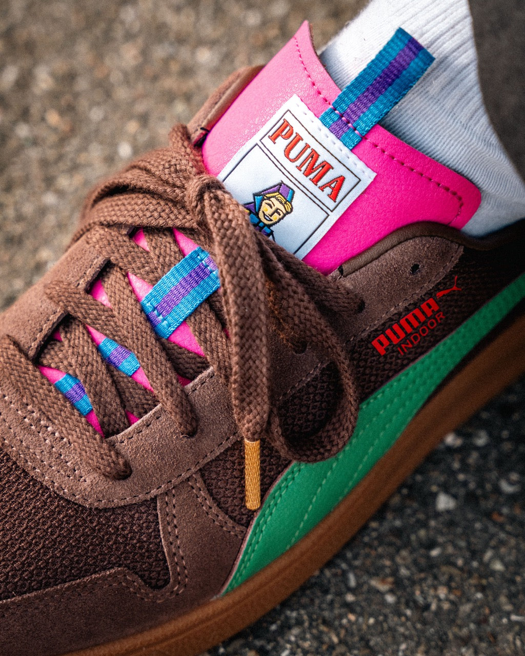 Eine Nahaufnahme des Puma Rothaus Indoor Tannenzäpfle Sneakers mit braunem Wildleder, grünen und rosa Akzenten, blau-gestreiften Schnürsenkeln und dem Puma Logo auf der Zunge, gepaart mit einer weißen Socke auf Beton. Inspiriert von Schwarzwälder Kultbier Vibes.