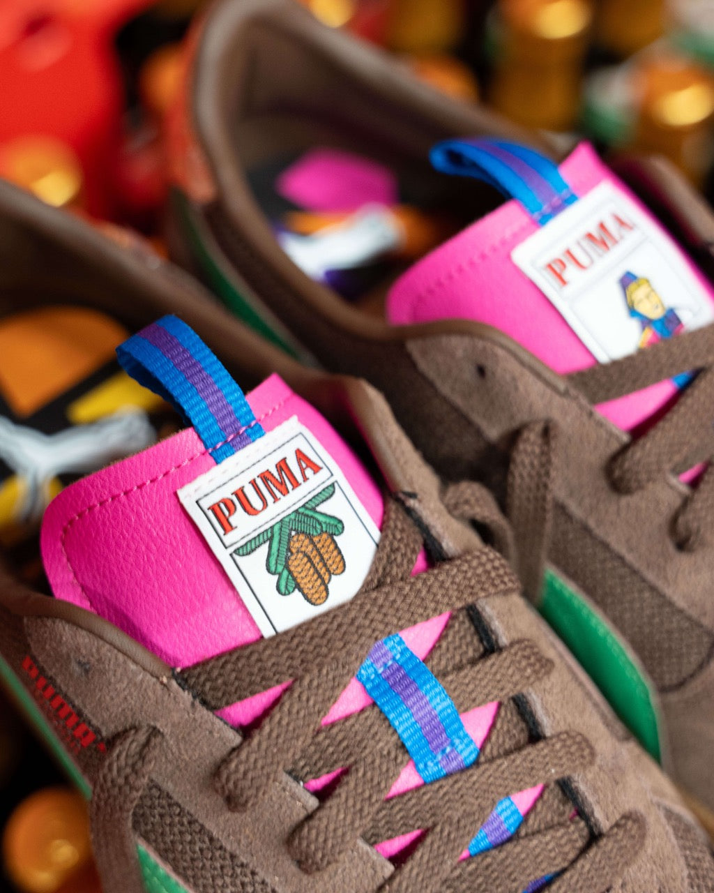 Nahaufnahme der braunen Rothaus Indoor Tannenzäpfle von Puma mit grünen Akzenten, rosa Zungen, blau und lila gestreiften Zuglaschen und einem einzigartigen Logo, das von den kultigen Tannenzäpfle Schwarzwälder Kultbier inspiriert ist.