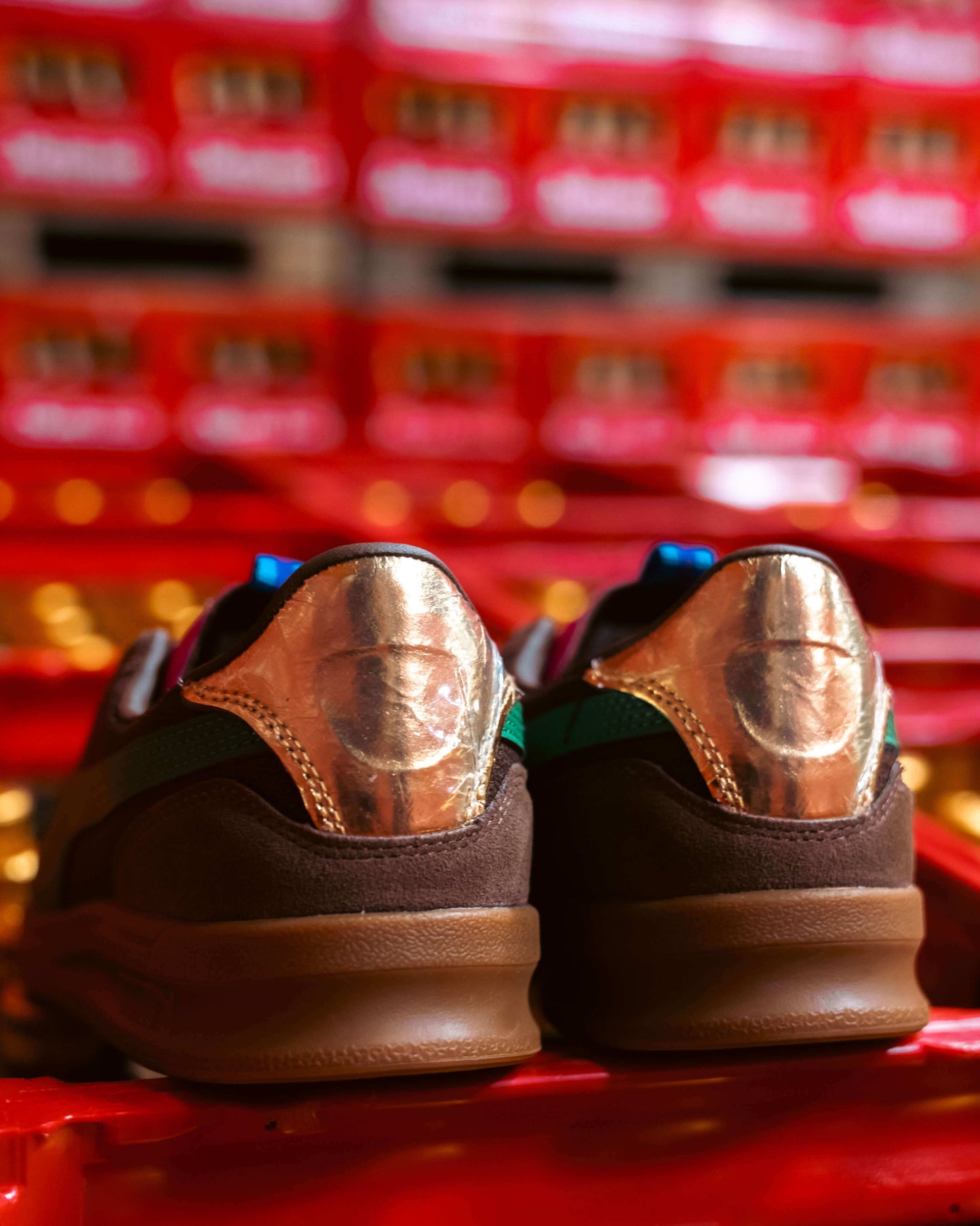 Eine Nahaufnahme der Rückseite von zwei braunen Puma Rothaus Indoor Tannenzäpfle Turnschuhen mit glänzenden kupferfarbenen Fersenkappen und geprägtem Gesicht, vor einem Hintergrund von gestapelten roten Tannenzäpfle-Boxen, leicht unscharf.