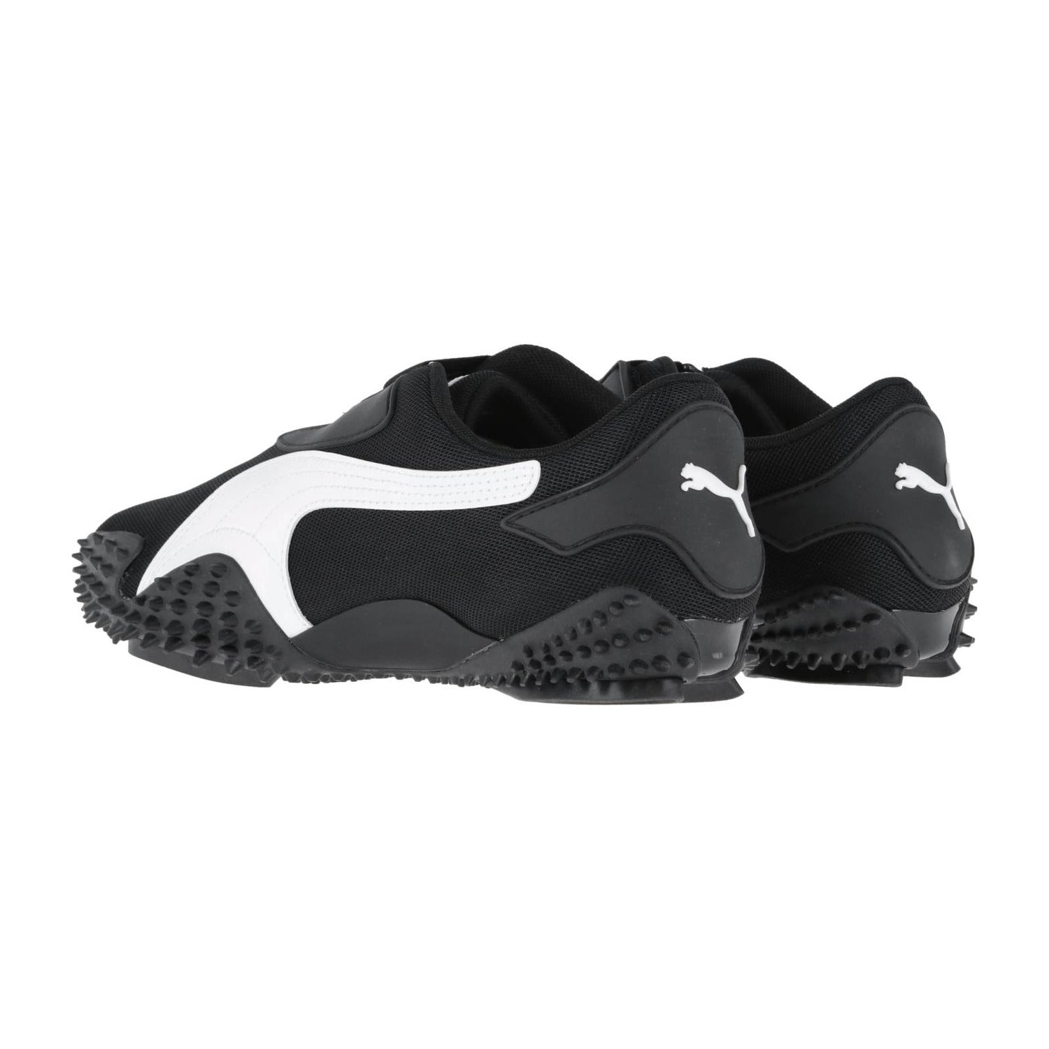 Ein Paar schwarze Puma Mostro OG Prime Turnschuhe mit weißen Puma Logos und Streifen, mit strukturierten Gummisohlen mit markanten Spikes für Grip, von hinten und von der Seite gezeigt.
