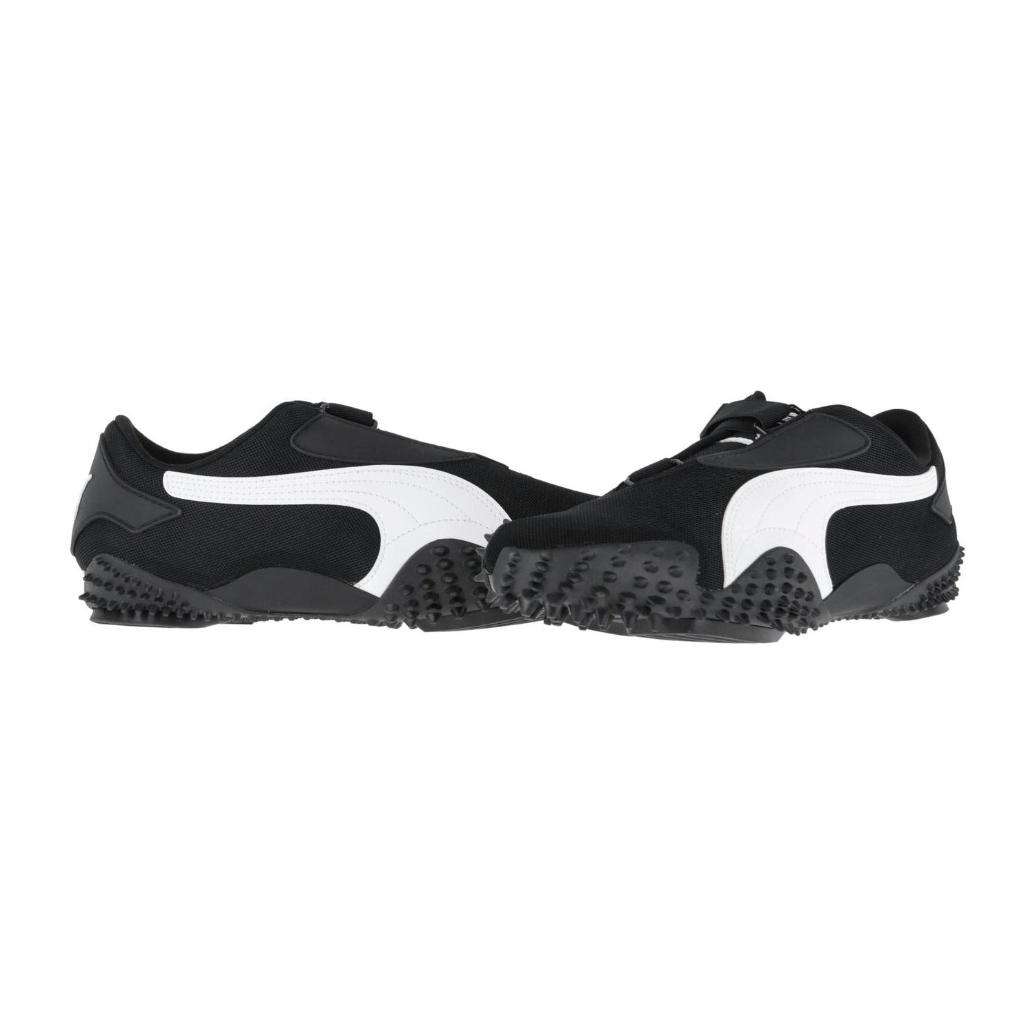 Ein Paar Puma Mostro OG Prime Sneakers in Schwarz, mit markanten weißen Seitenstreifen und strukturierten, gummierten Sohlen, stehen sich auf weißem Hintergrund gegenüber.