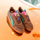 Die Rothaus Indoor Tannenzäpfle Sneakers von Puma haben ein braunes Obermaterial, blaugrüne Seitenstreifen, pinke und blaugrüne Akzente und mehrfarbige Schnürsenkel, die auf einer leuchtend orangefarbenen Oberfläche präsentiert werden und von Tannenzäpfle Kultbier inspiriert sind.