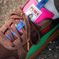 Eine Nahaufnahme des Puma Rothaus Indoor Tannenzäpfle Sneakers mit braunem Wildleder, grünen und rosa Akzenten, blau-gestreiften Schnürsenkeln und dem Puma Logo auf der Zunge, gepaart mit einer weißen Socke auf Beton. Inspiriert von Schwarzwälder Kultbier Vibes.