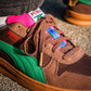 Eine Nahaufnahme des Puma Rothaus Indoor Tannenzäpfle Sneakers mit braunem Wildleder, grünem Streifen, rosa und blauen Zungenakzenten und goldenen Schnürsenkeln - eine Hommage an das Schwarzwälder Kultbier Tannenzäpfle - auf Beton getragen.