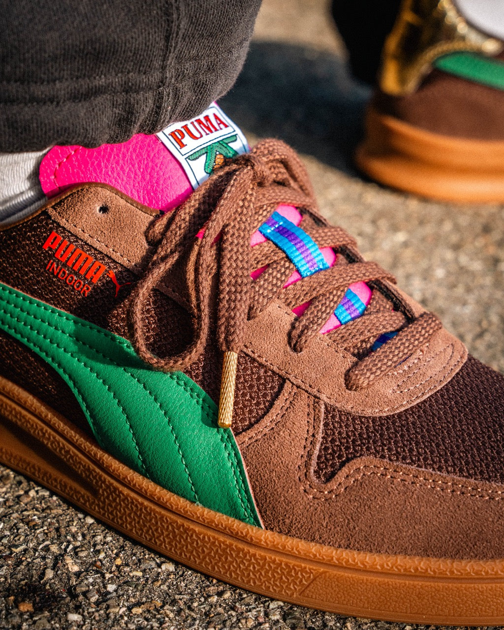 Eine Nahaufnahme des Puma Rothaus Indoor Tannenzäpfle Sneakers mit braunem Wildleder, grünem Streifen, rosa und blauen Zungenakzenten und goldenen Schnürsenkeln - eine Hommage an das Schwarzwälder Kultbier Tannenzäpfle - auf Beton getragen.