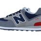 Der New Balance 574 V2 Sneaker in Grau, Marineblau und Weiß hat ein großes N-Logo, rote Fersenakzente, eine ENCAP-Zwischensohle für Komfort und eine dunkelgraue Laufsohle. Abgebildet im Seitenprofil vor einem weißen Hintergrund.