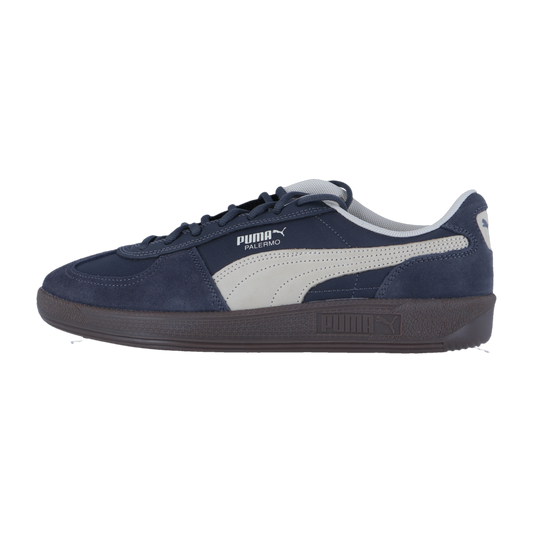 Der Puma Palermo Pop Retro-Sneaker hat ein marineblaues und weißes Wildleder-Obermaterial mit dem Puma-Logo und einer dunklen Sohle, die vor einem schlichten weißen Hintergrund steht.