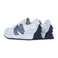 Die New Balance WS327V1 Sneakers haben ein weißes und graues Design, dunkelblaue Sohlen und ein großes blaues N-Logo an den Seiten, das von hinten schräg zu sehen ist.