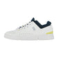 Der On THE ROGER ADVANTAGE ist ein weißer Low-Top-Sneaker mit dunkelblauem Futter, gelbem Fersenakzent, perforierten Seiten und einem grauen On-Logo. Er hat weiße Schnürsenkel und eine dicke weiße Sohle für klassischen Stil und Komfort.