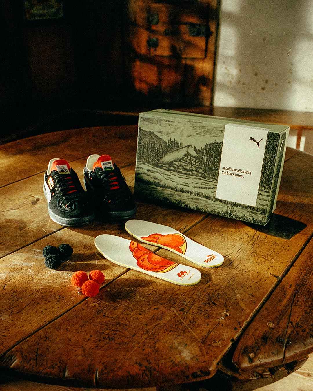 Ein Paar schwarze Puma-Sneaker, ein Schuhkarton mit Waldkunst, bunte Einlegesohlen mit Tortenstückchenmuster und rote und schwarze Bommeln sind auf einem rustikalen Holztisch in warmem, natürlichem Licht arrangiert.