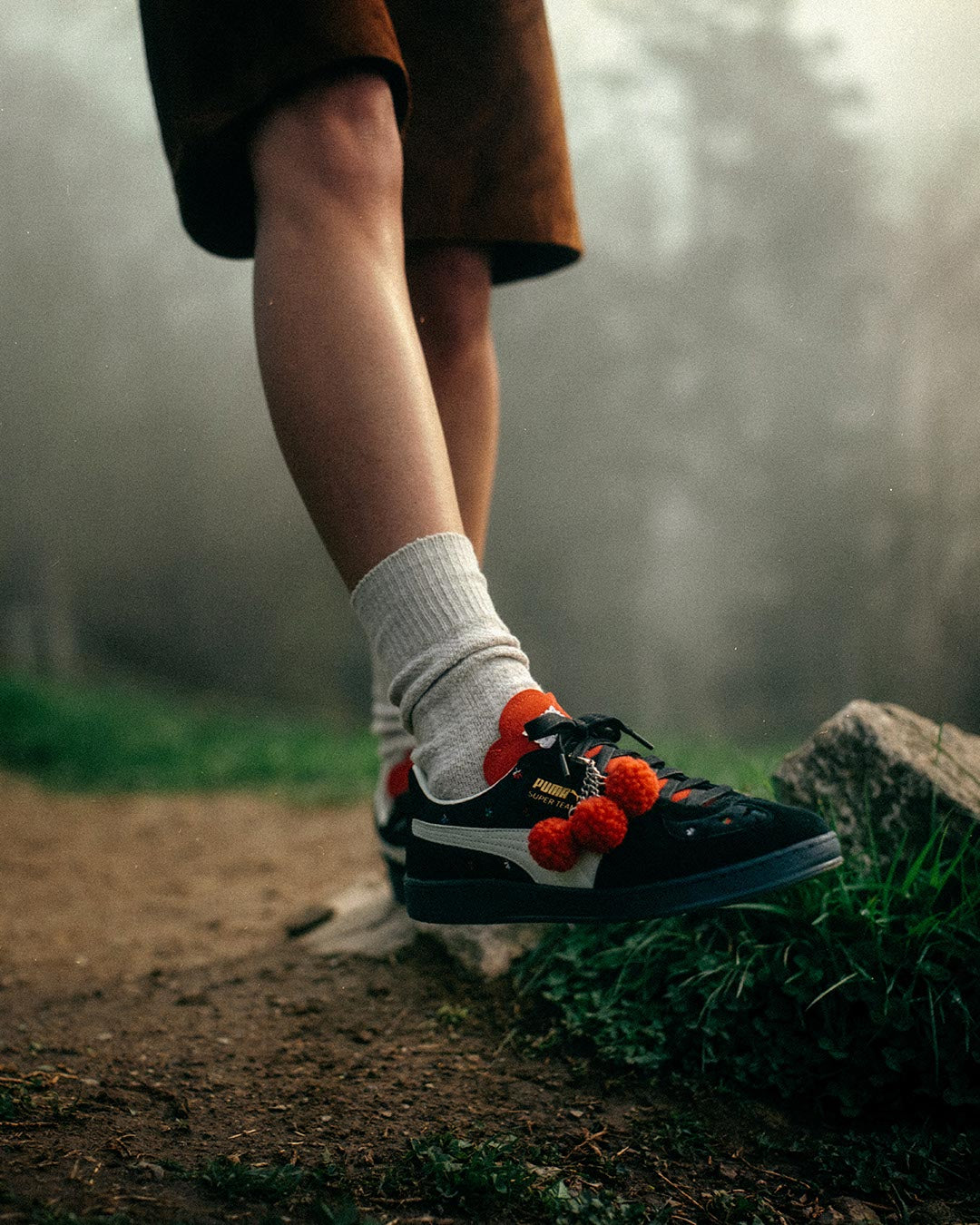 Eine Person steht auf einem unbefestigten Waldweg und trägt eine weiße Socke und einen schwarzen Turnschuh mit leuchtend roten Bommeln. Der Hintergrund ist neblig und grün, und ein Fuß ruht auf einem Felsen.