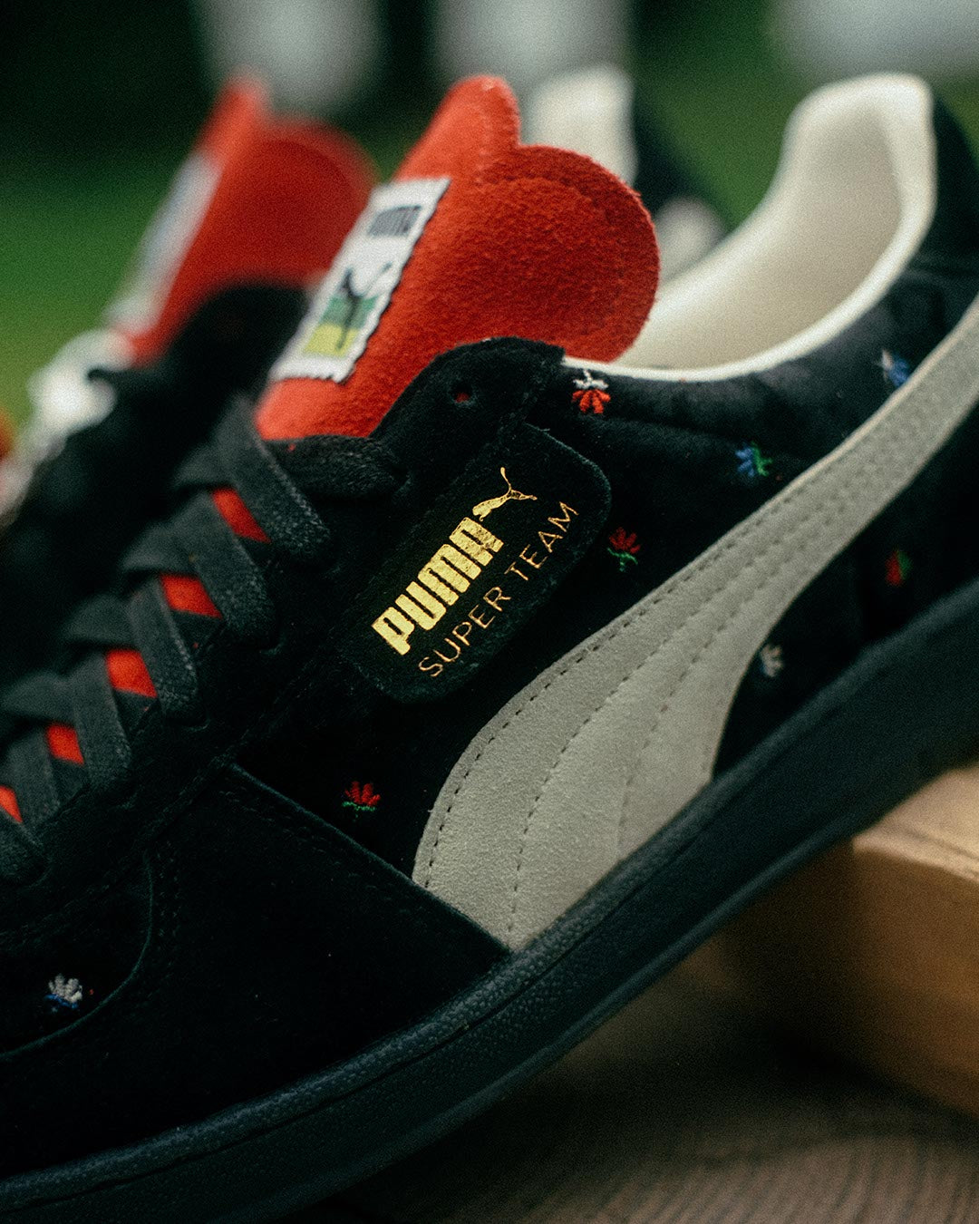 Nahaufnahme eines schwarzen Puma Super Team Sneakers mit weißem Streifen, roten Akzenten auf der Zunge und den Schnürsenkeln sowie kleinen mehrfarbigen Stickereien auf dem Obermaterial. Das goldene Puma-Logo ist an der Seite sichtbar.